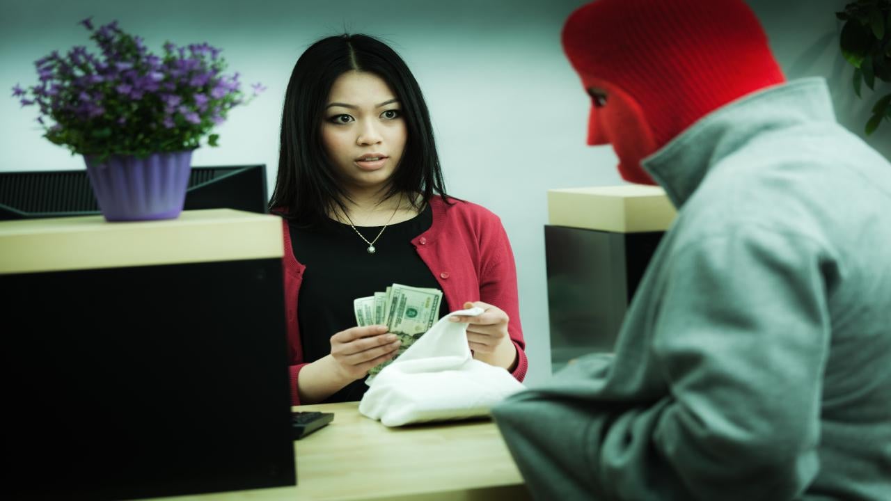 A young, frightened female bank teller places a large wad of cash in a sack as she looks at a bank robber concealed in a red mask, pointing at her in his hoodie pocket.