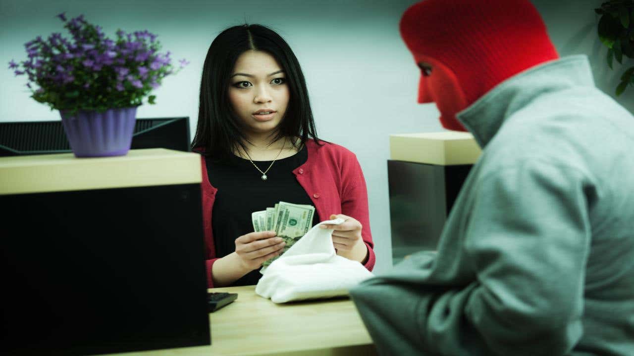 A young, frightened female bank teller places a large wad of cash in a sack as she looks at a bank robber concealed in a red mask, pointing at her in his hoodie pocket.