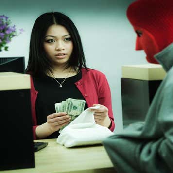 A young, frightened female bank teller places a large wad of cash in a sack as she looks at a bank robber concealed in a red mask, pointing at her in his hoodie pocket.