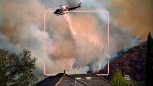 helicopter flying above wildfire in smoke-filled sky spraying water