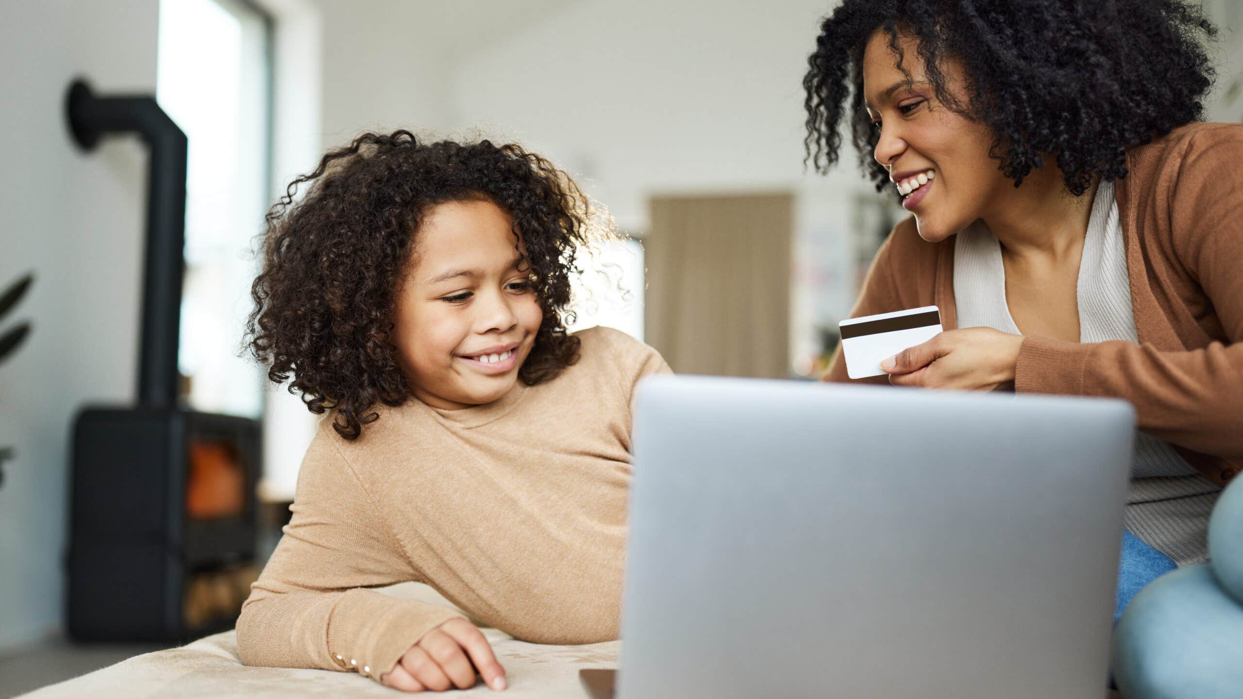 Happy African American single mother and daughter using credit card while shopping online over laptop at home.