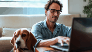 Middle-aged man comfortably working at home on his laptop, sitting on the floor with his Beagle dog by his side.