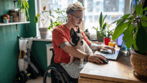 Man working on laptop, sitting in wheelchair with small dog on his lap.