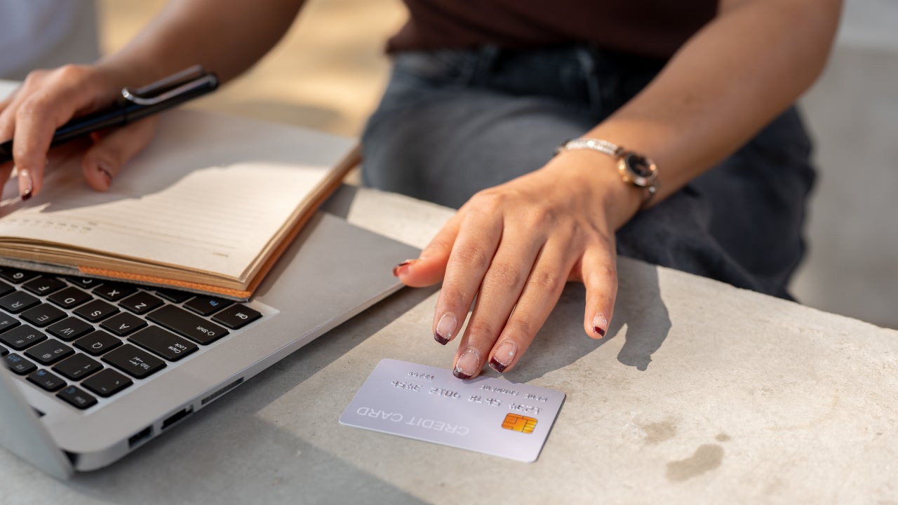 A close-up of a woman's hand holds a credit card above a table while using her laptop.