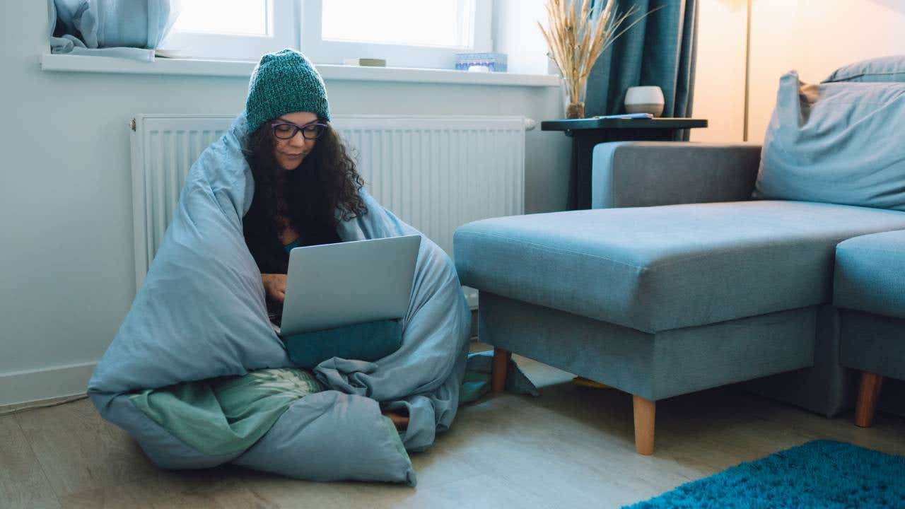 A woman sits on the floor, bundled up in a comforter with her laptop.