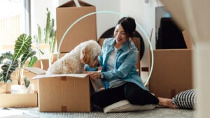 Image of a woman sitting among packing boxes. The box next to her is open and has a golden doodle dog sitting inside of it.