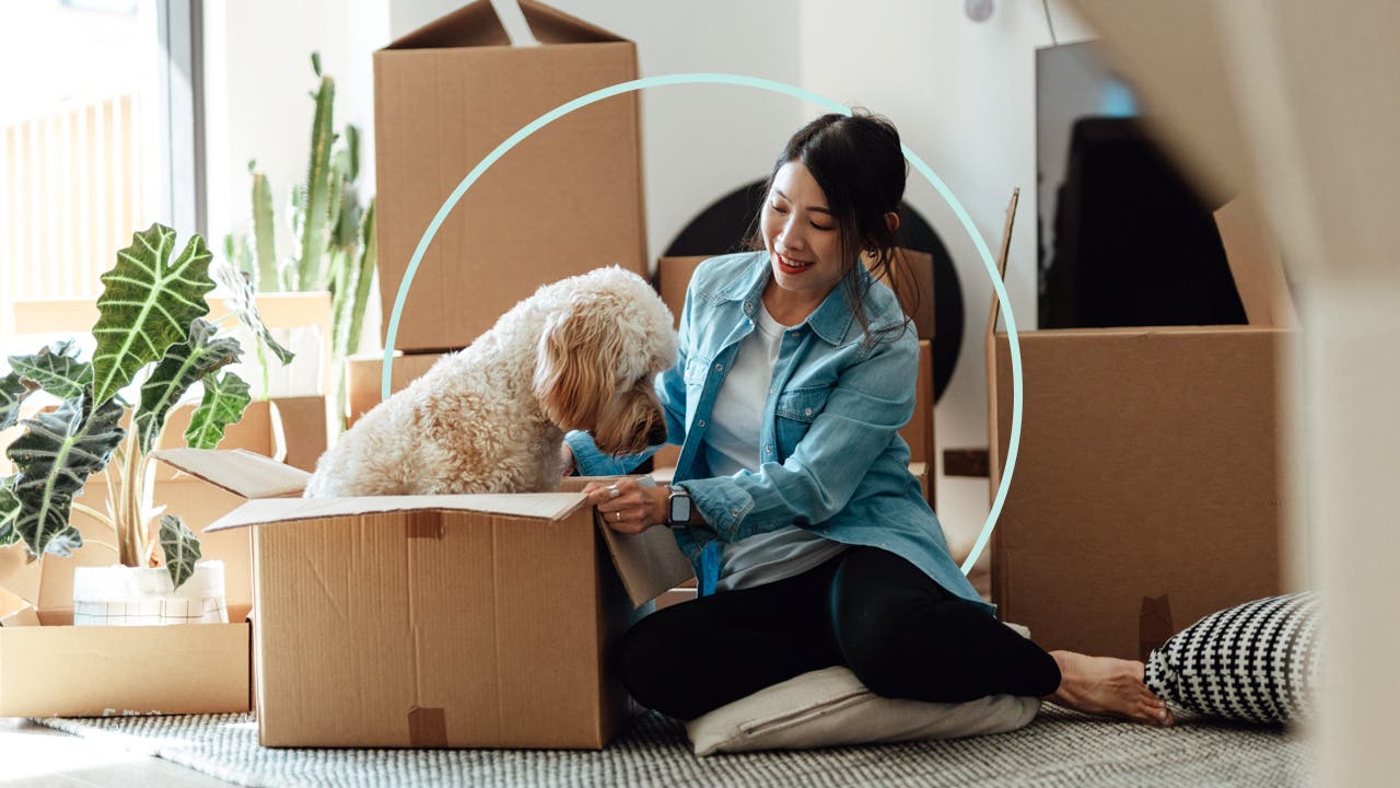 Image of a woman sitting among packing boxes. The box next to her is open and has a golden doodle dog sitting inside of it.