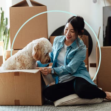 Image of a woman sitting among packing boxes. The box next to her is open and has a golden doodle dog sitting inside of it.