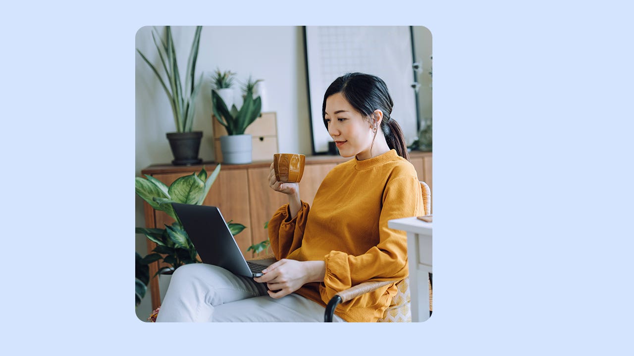 Woman sitting in a chair with her laptop open on her lap. She is holding a mug in her right hand.