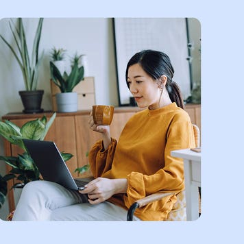 Woman sitting in a chair with her laptop open on her lap. She is holding a mug in her right hand.