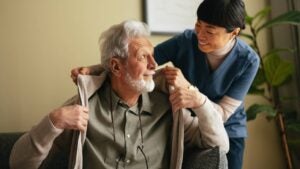 Close up shot of a smiling elderly man sitting on the sofa and a his caregiver. She is smiling while helping him put on or take off his jacket.