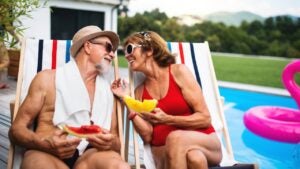 Cheerful senior couple sitting by swimming pool outdoors in backyard, talking.