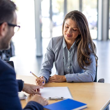 A white woman in business casual clothing signs paperwork at a car dealership. She sits across from a white man in a suit.