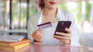 A cropped image of a young woman who sits at a table in a cafeteria holding a credit card and a smartphone