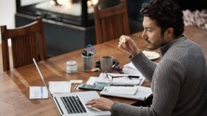 Younger man in his 30s paying bills on his computer, with calculator, papers, bills, coffee and a pair of glasses on the table by his side.