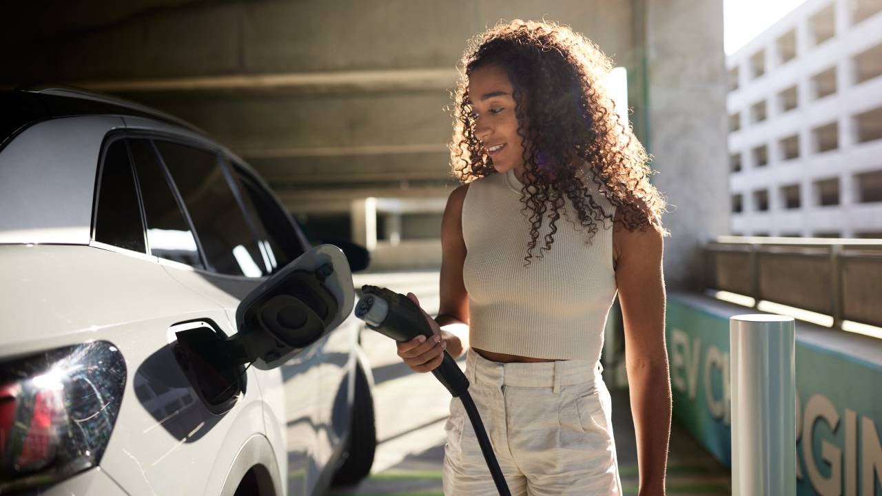 Young woman holding electric plug by car at charging station