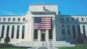 A wide-angle image of the front of the Federal Reserve HQ in Washington, DC. The U.S. flag is prominently displayed on the front of the building.