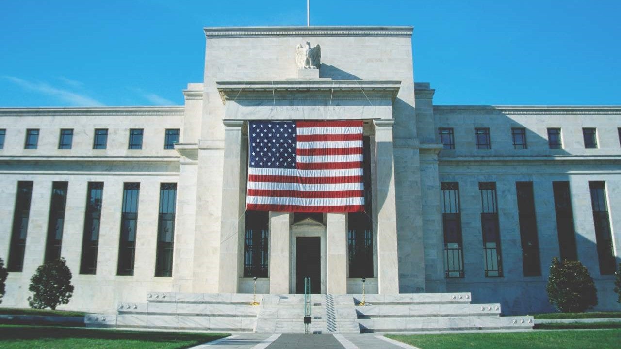 A wide-angle image of the front of the Federal Reserve HQ in Washington, DC. The U.S. flag is prominently displayed on the front of the building.