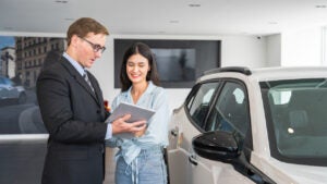 A male salesperson shows a young woman content on a tablet. They stand next to a silver-white SUV.