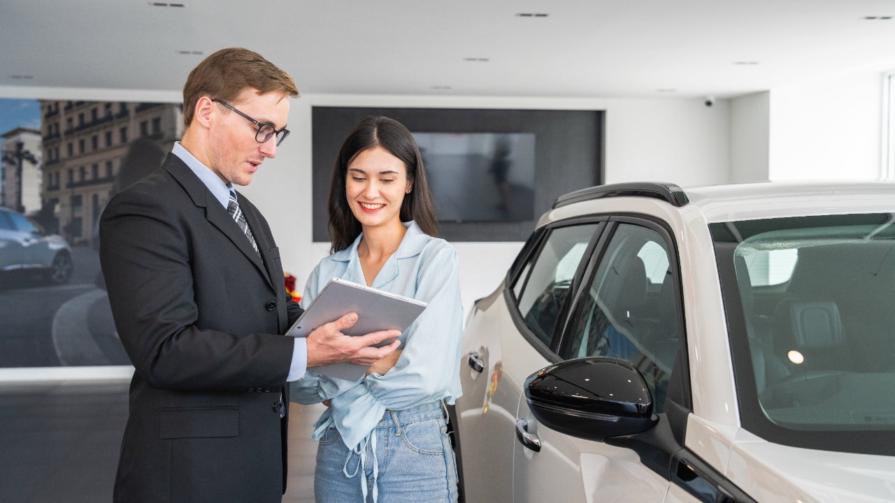 A male salesperson shows a young woman content on a tablet. They stand next to a silver-white SUV.