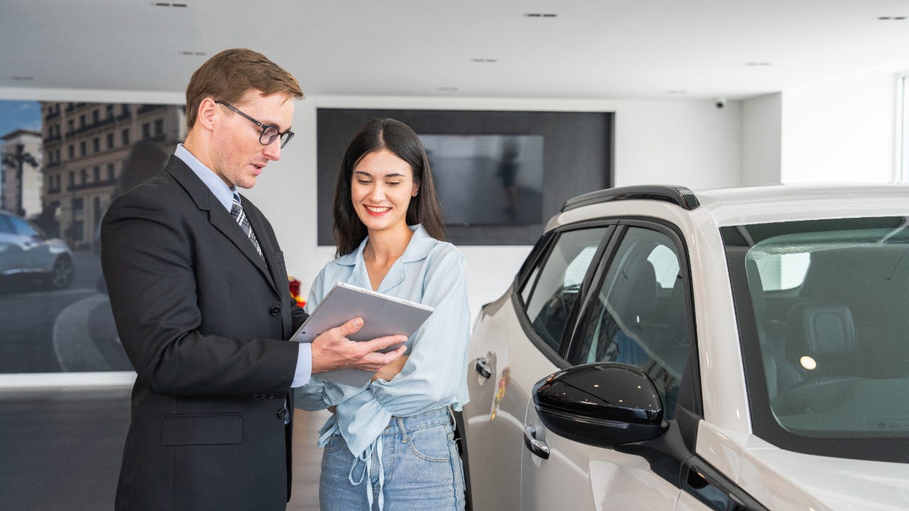 A male salesperson shows a young woman content on a tablet. They stand next to a silver-white SUV.