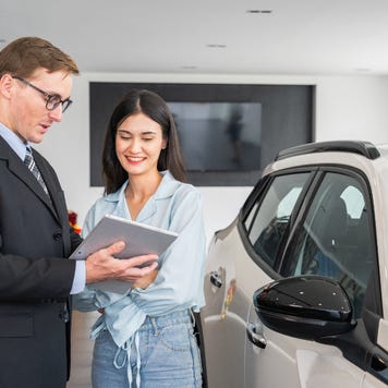 A male salesperson shows a young woman content on a tablet. They stand next to a silver-white SUV.