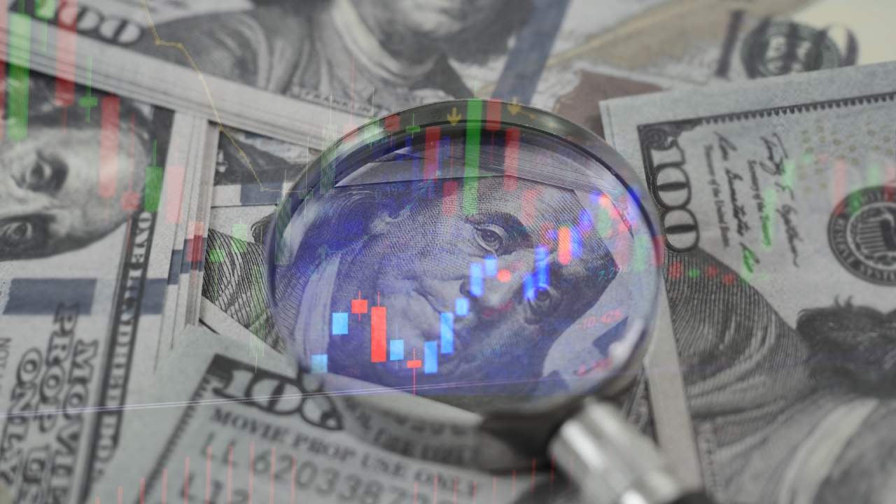 A magnifying glass on top of a $100, magnifying Benjamin Franklin's face, and surrounded by other $100 bills.