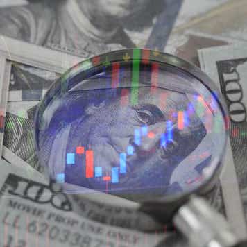 A magnifying glass on top of a $100, magnifying Benjamin Franklin's face, and surrounded by other $100 bills.