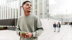 A young Black university student on his phone smiles and looks up to the right.