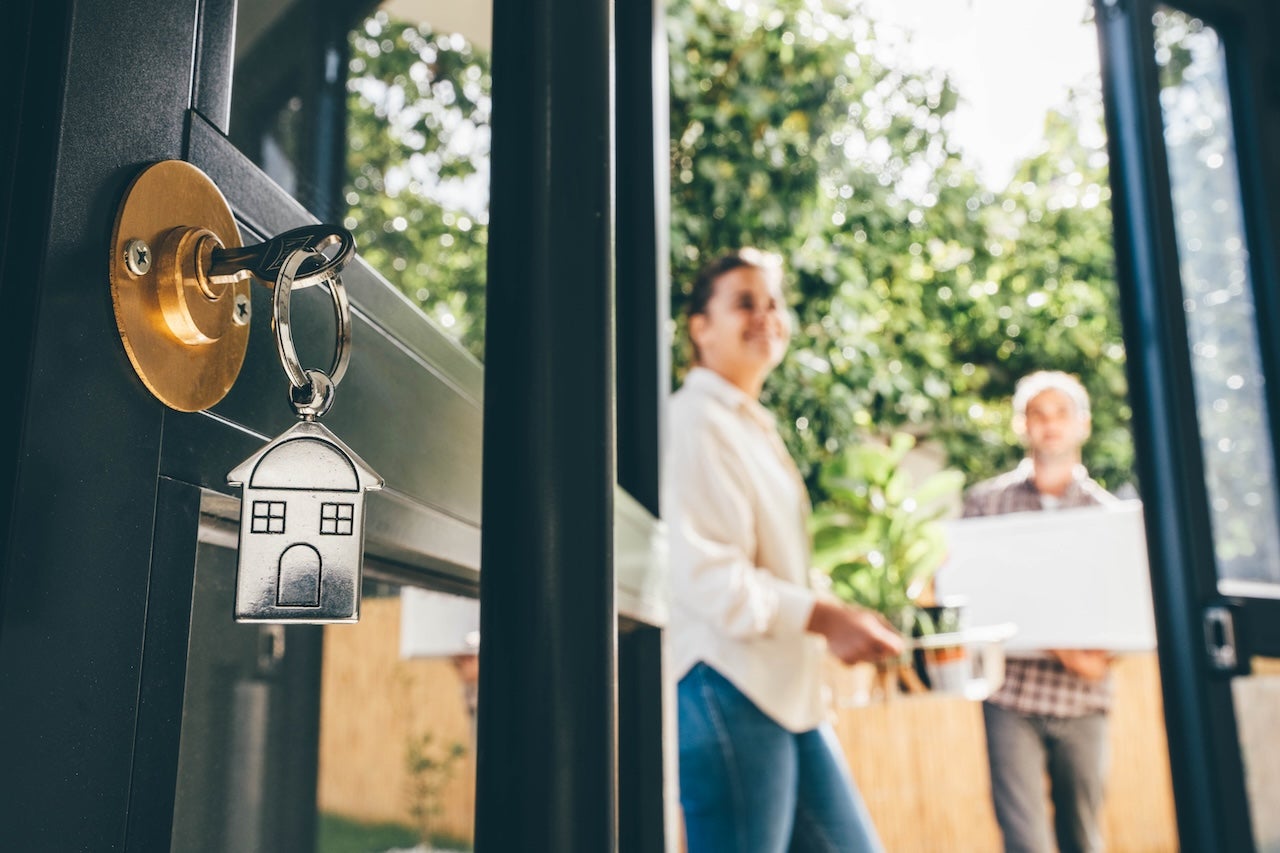 Keys in a door lock with couple celebrating moving day in the background