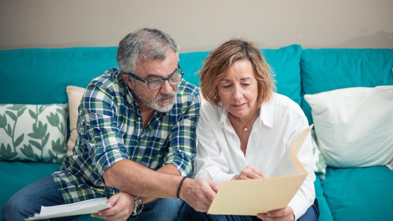 An older couple sits on a blue couch and reviews financial documents.