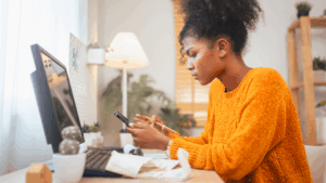 A woman in a yellow sweater is focused on her smartphone, with a computer monitor and notepad on the desk in a bright room.