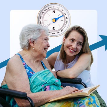 Image of an older woman sitting in a wheelchair reading a book with a younger women squatting down next to her. There is a large compass above and behind them .