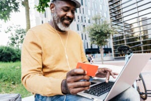 Smiling man sites on a park bench holding a credit card while looking at his laptop.