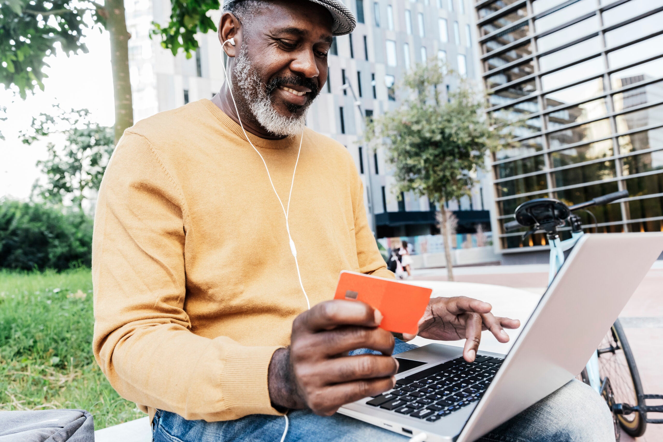 Smiling man sites on a park bench holding a credit card while looking at his laptop.