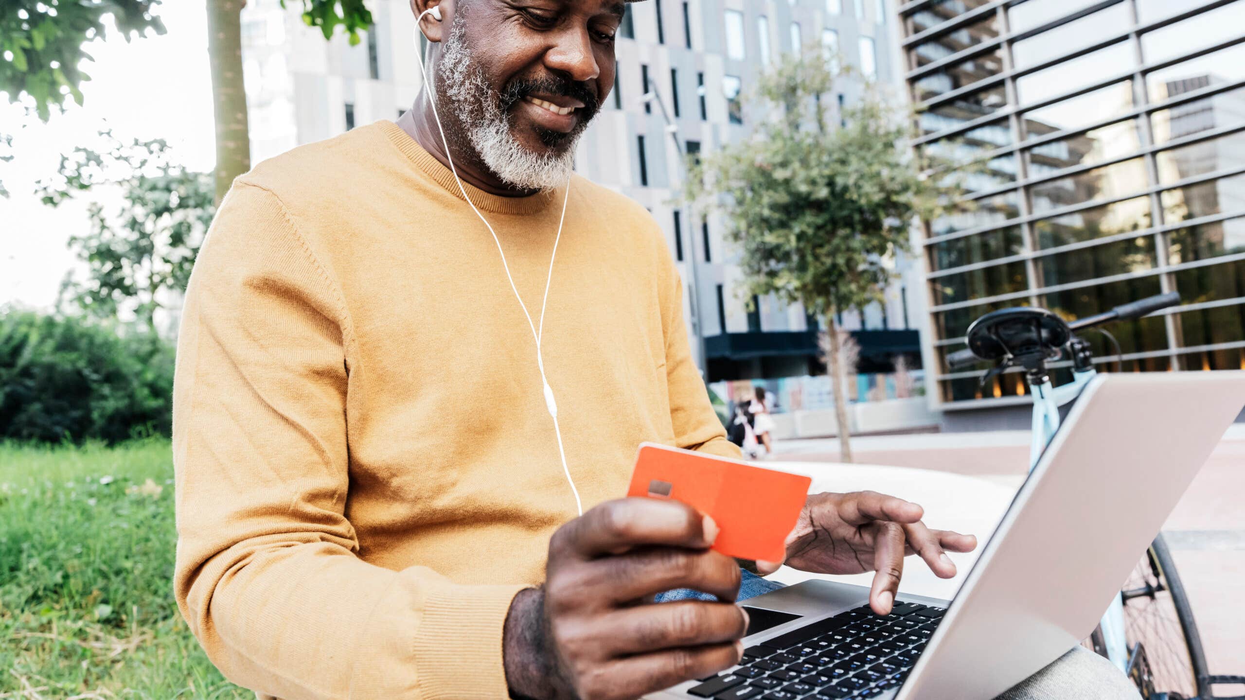 Smiling man sites on a park bench holding a credit card while looking at his laptop.