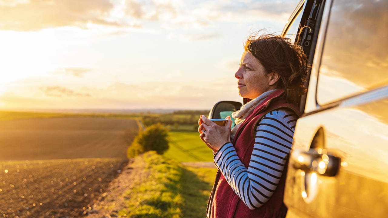 A woman (in her 40s) takes in the view of the countryside from her campervan. She looks contented and relaxed.