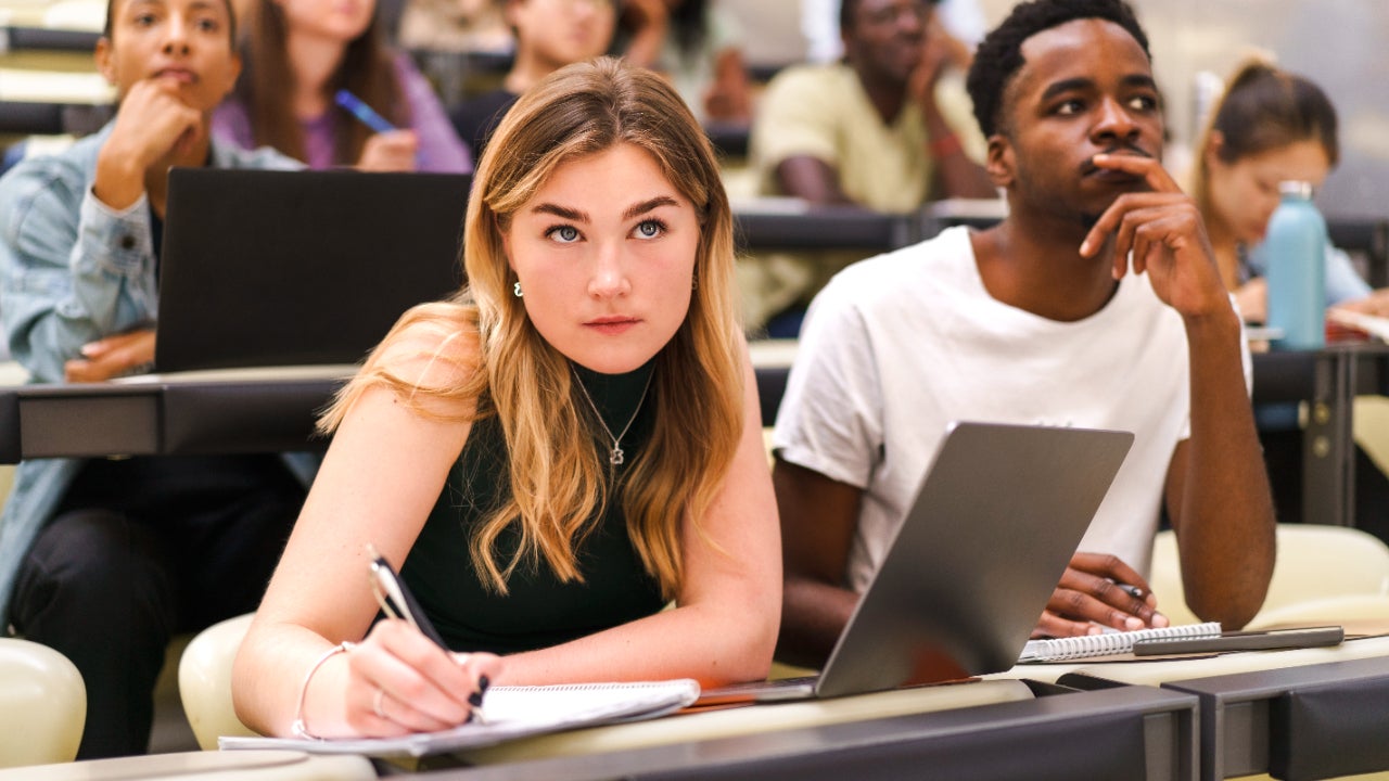 Picture of university lecture hall focused on a young pale woman taking notes in a notebook and a young Black man looking inquisitive.