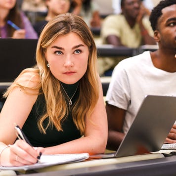 Picture of university lecture hall focused on a young pale woman taking notes in a notebook and a young Black man looking inquisitive.