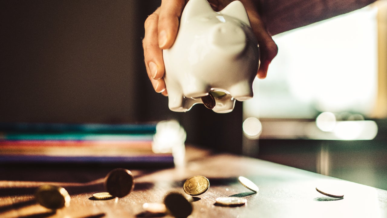 A hand holding a white piggy bank. There is a table with a variety of coins underneath.