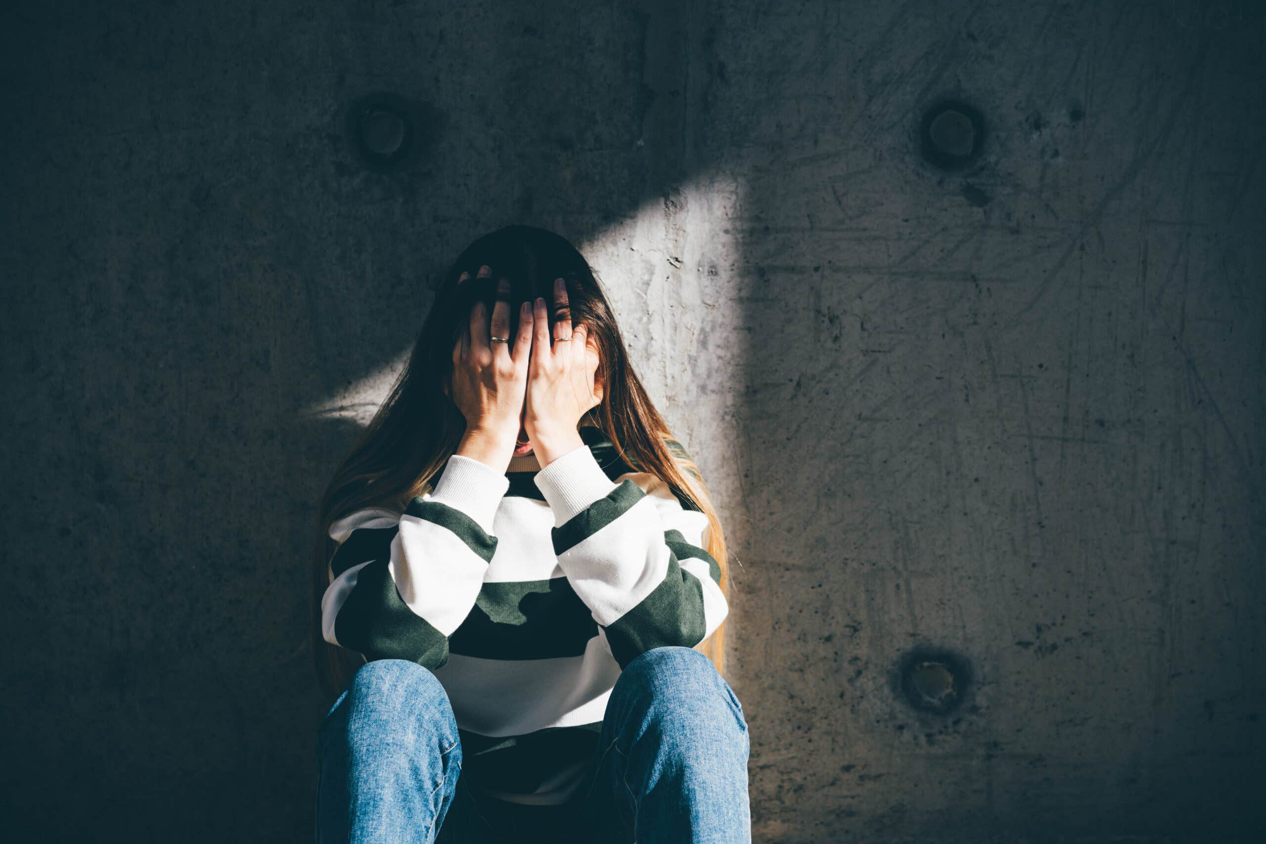 Woman sits against a concrete wall hiding her face in her hands