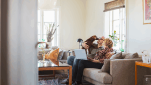 A couple sitting on their sofa in the living room, with a laptop and their dog.