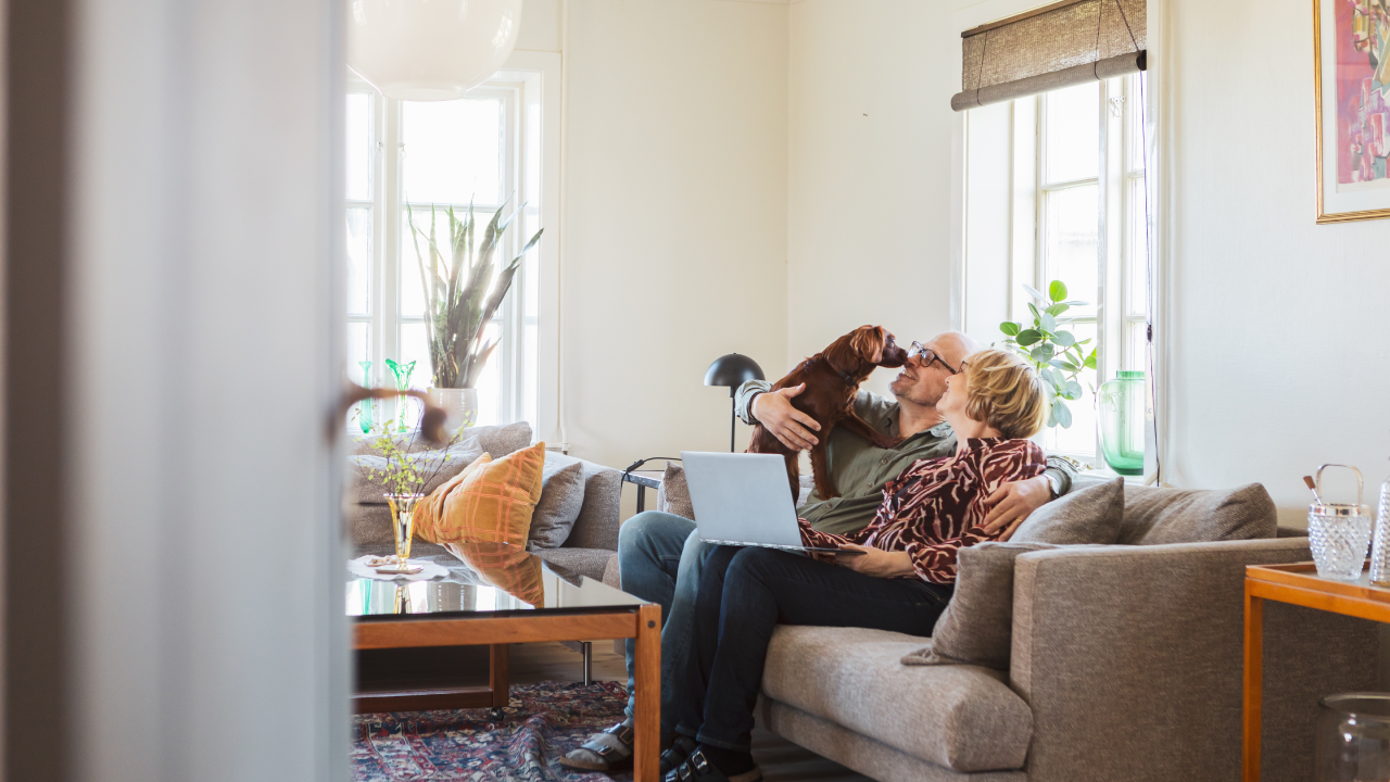 A couple sitting on their sofa in the living room, with a laptop and their dog.