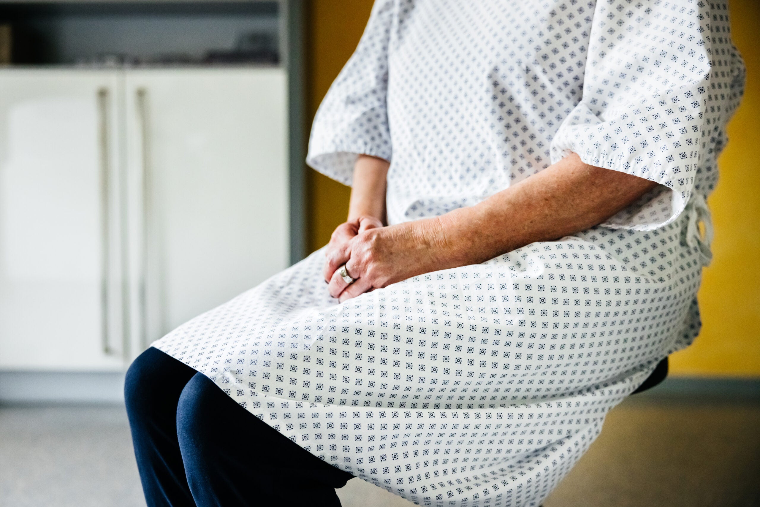 A person waits in a hospital gown with hands folded on their lap.