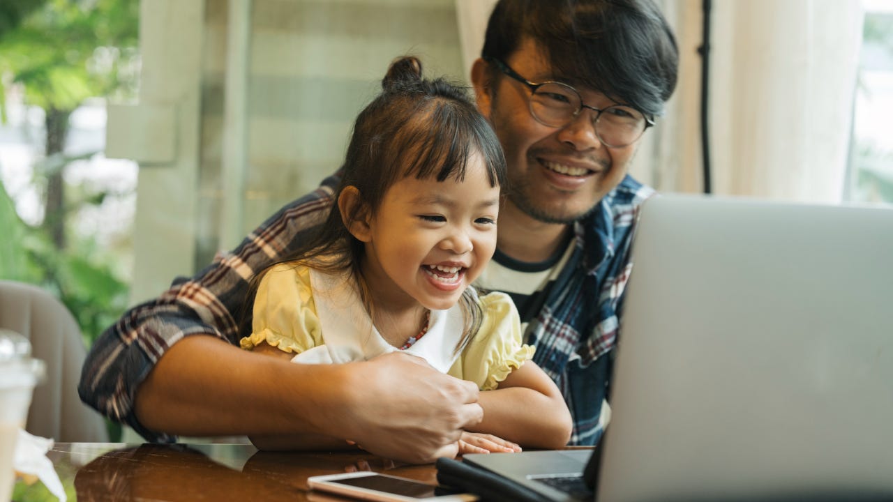 Smiling father holds smiling daughter on his lap while looking at a computer