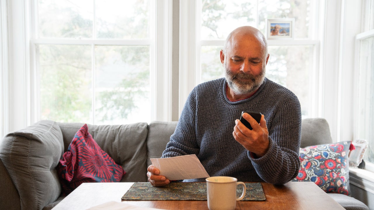 A man sits on a couch while reading his phone and reviewing a piece of mail.