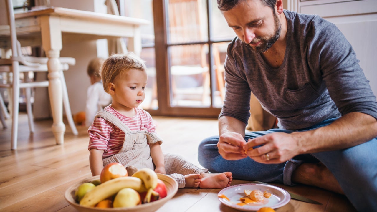 Mature father with small toddler daughter sitting on the floor indoors, eating fruit.